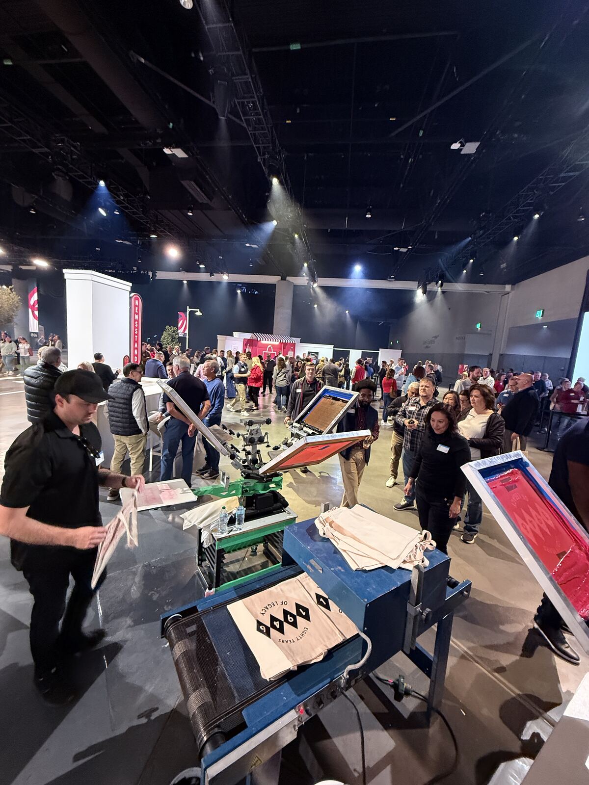Large nighttime live event with dozens of attendees, product displays in protective cases, and stage lighting